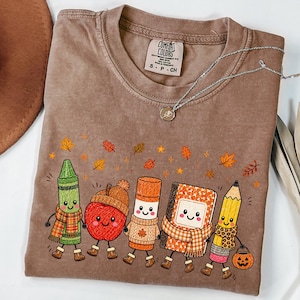 May include: A brown Comfort Colors t-shirt featuring a graphic of a green crayon, a red apple with an acorn hat, an orange glue stick, a notebook, and a yellow pencil, all with faces and autumn accessories, surrounded by falling orange and yellow leaves.