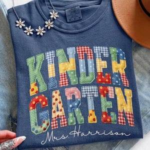 May include: A blue crewneck t-shirt featuring the word KINDERGARTEN in large, multi-colored, patterned block letters with a patchwork aesthetic. Below the word, the text Mrs. Harrison is printed in white script. A silver floral necklace is visible.