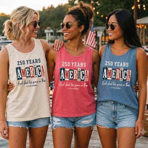 Könnte beinhalten: Drei ärmellose Tanktops in Creme, Pink und Blau, jeweils mit dem Text 250 YEARS AMERICA, God shed his grace on thee und 1776-2026. Die Grafik enthält ein stilisiertes amerikanisches Flaggendesign mit Sternen und Streifen in Rot, Weiß und Blau.