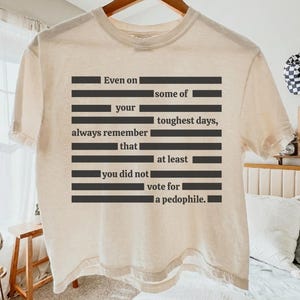 Può includere: T-shirt color crema con un design testuale nero. Il testo recita: "Anche in alcuni dei tuoi giorni più difficili, ricorda sempre che non hai votato per un pedofilo."