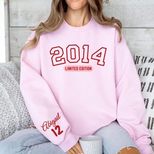 May include: A light pink sweatshirt with the year "2014" in red block letters, with "LIMITED EDITION" below. The name "Abigail" and the number "12" are printed on the left sleeve in red script. The sweatshirt is a crew neck style.