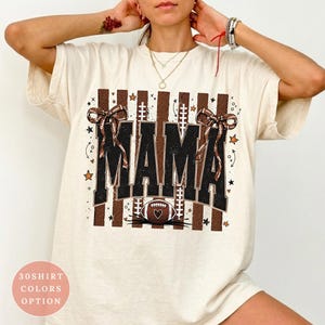 以下が含まれることがあります： アイボリーの半袖Tシャツ。黒いブロック体で「MAMA」と書かれたグラフィックプリントが特徴です。茶色の縦縞、2つの茶色の柄入りリボン、ハート付きのフットボールのアイコンがデザインされています。テキストは「COMFORT COLORS」と記載されています。