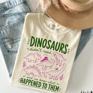 May include: Ivory-colored t-shirt with the text "DINOSAURS didn't read and we know what HAPPENED TO THEM" in green and pink. The shirt features dinosaur illustrations. The shirt is paired with light blue jeans and a tan hat.