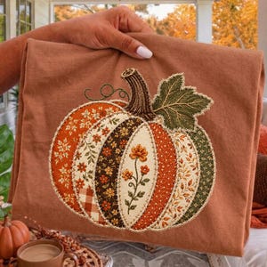 May include: A rust-colored t-shirt featuring a large, embroidered pumpkin graphic. The pumpkin is composed of vertical fabric panels in various autumn-themed patterns, including floral, gingham, and solid textures, finished with a green leaf.