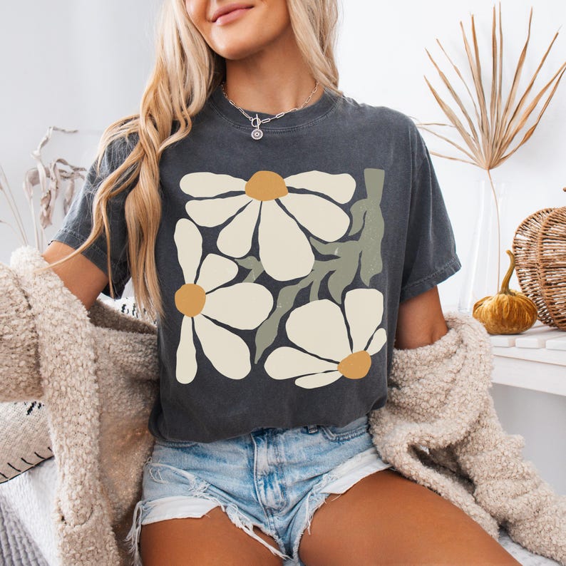 Puede incluir: Camiseta gris oscuro con un dise&ntilde;o floral de grandes margaritas blancas, centros amarillos y tallos verdes. La camiseta est&aacute; hecha de material suave y es adecuada para ropa casual. La camiseta se combina con pantalones cortos de mezclilla.