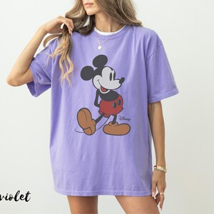 May include: A violet-colored t-shirt featuring a classic Mickey Mouse graphic. The cartoon character is depicted in his iconic pose, wearing red shorts and brown shoes. The Disney logo is visible. The word "violet" is printed in black at the bottom left.