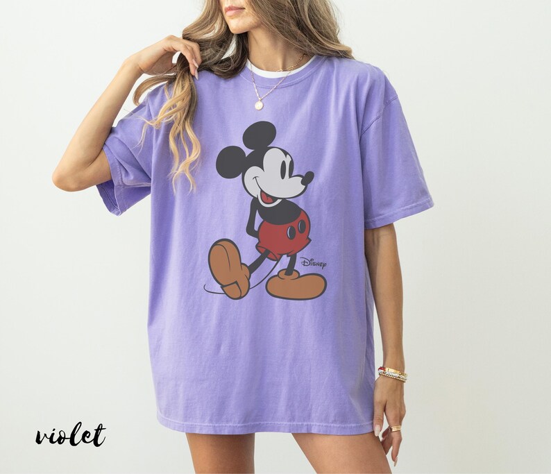 May include: A violet-colored t-shirt featuring a classic Mickey Mouse graphic. The cartoon character is depicted in his iconic pose, wearing red shorts and brown shoes. The Disney logo is visible. The word "violet" is printed in black at the bottom left.