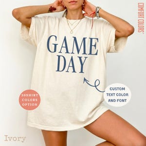 May include: An ivory short-sleeve t-shirt featuring the dark blue text GAME DAY printed on the front. The shirt includes a Comfort Colors label, a circular graphic noting 30 shirt colors, and a circular graphic stating custom text color and font.