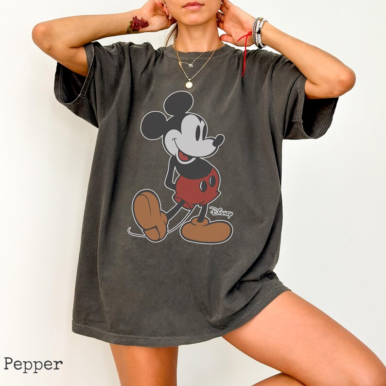 May include: Pepper-colored oversized t-shirt featuring a classic Mickey Mouse graphic. The cartoon character is depicted in his iconic red shorts, brown shoes, and white gloves. The Disney logo is visible below the graphic.