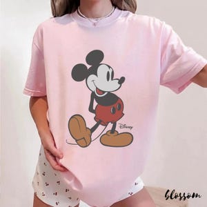 May include: A light pink t-shirt featuring a classic Mickey Mouse graphic. The cartoon character is depicted in black, red, and tan, with the Disney logo. The word "Blossom" is printed in a script font at the bottom right.