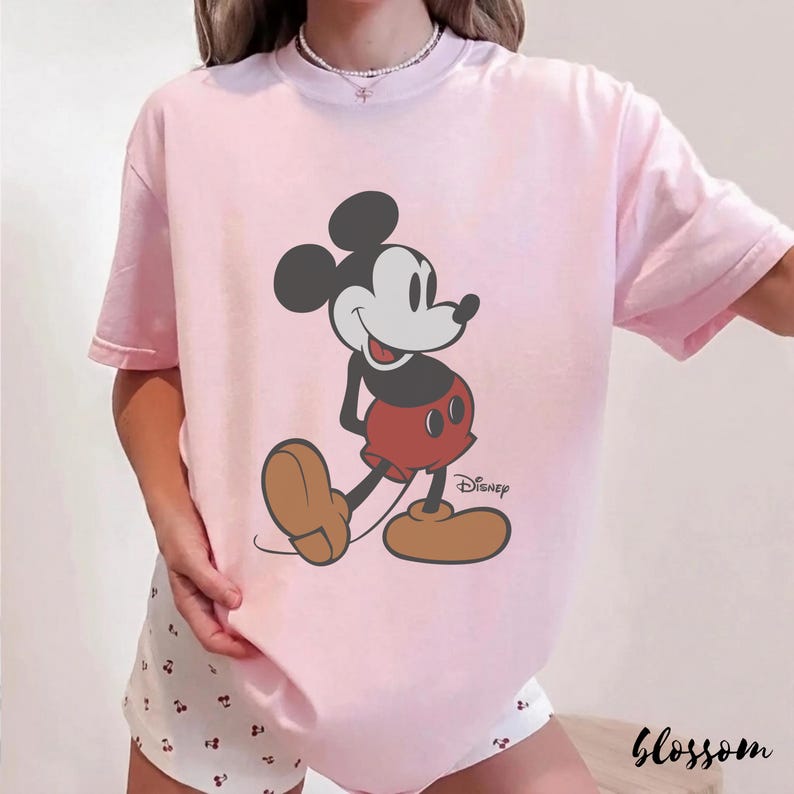 May include: A light pink t-shirt featuring a classic Mickey Mouse graphic. The cartoon character is depicted in black, red, and tan, with the Disney logo. The word "Blossom" is printed in a script font at the bottom right.