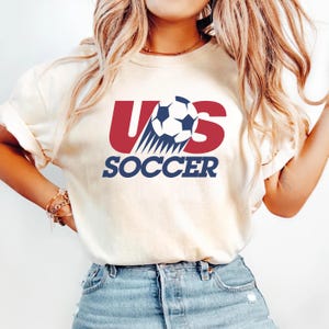May include: A cream-colored short-sleeve t-shirt featuring a graphic design with the red letters U and S, a central black and white soccer ball, and the word SOCCER printed in bold blue capital letters underneath the graphic.