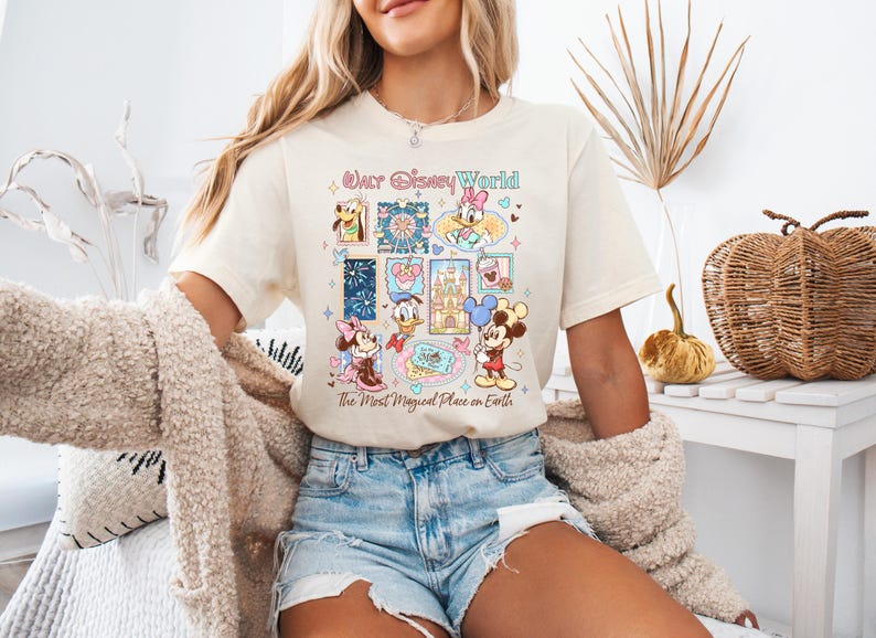 K&ouml;nnte beinhalten: Cremefarbenes T-Shirt mit einer farbenfrohen Grafik von Disney-Figuren und -Attraktionen. Das Design umfasst Micky und Minnie Maus, Donald Duck und andere Charaktere. Der Text auf dem Shirt lautet "Walt Disney World" und "The Most Magical Place on Earth".