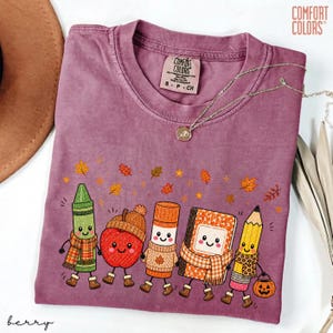May include: A berry-colored short-sleeve t-shirt with a Comfort Colors label at the neck. The graphic features five anthropomorphic school supplies including a crayon, apple, glue stick, notebook, and pencil, all wearing scarves amidst falling autumn leaves.