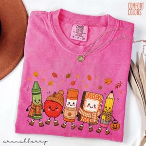 May include: A pink short-sleeve t-shirt featuring a graphic of a green crayon, a red apple, an orange glue stick, a notebook, and a yellow pencil, all with faces and scarves. The shirt has a Comfort Colors label at the neck and autumn leaves above the characters.