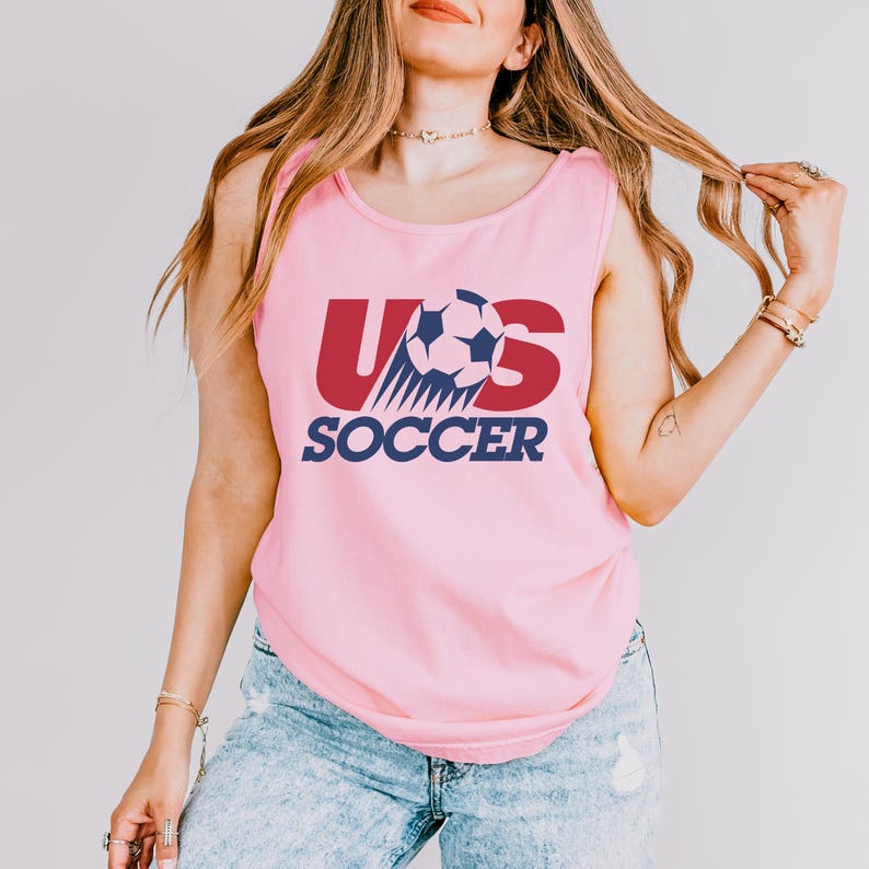 May include: A light pink sleeveless tank top featuring a graphic design with the text US SOCCER in dark blue and red. The letter U and S are in red, while the word SOCCER is in dark blue. A soccer ball graphic is positioned between the letters.