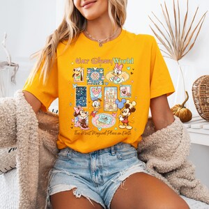 K&ouml;nnte beinhalten: Senfgelbes T-Shirt mit einer farbenfrohen Grafik von Disney-Figuren und -Attraktionen. Das Design umfasst Micky und Minnie Maus, Donald Duck und den Text "Our Disney World" und "The Most Magical Place on Earth."