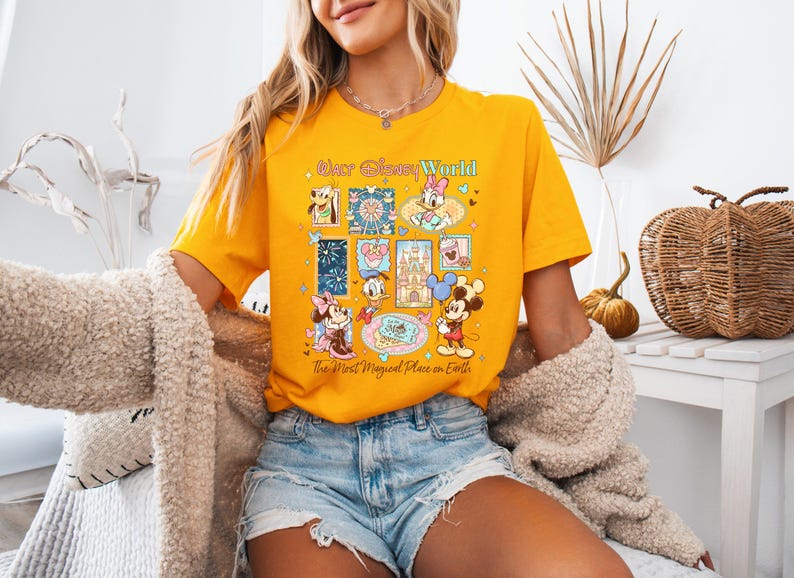 K&ouml;nnte beinhalten: Senfgelbes T-Shirt mit einer farbenfrohen Grafik von Disney-Figuren und -Attraktionen. Das Design umfasst Micky und Minnie Maus, Donald Duck und den Text "Our Disney World" und "The Most Magical Place on Earth."