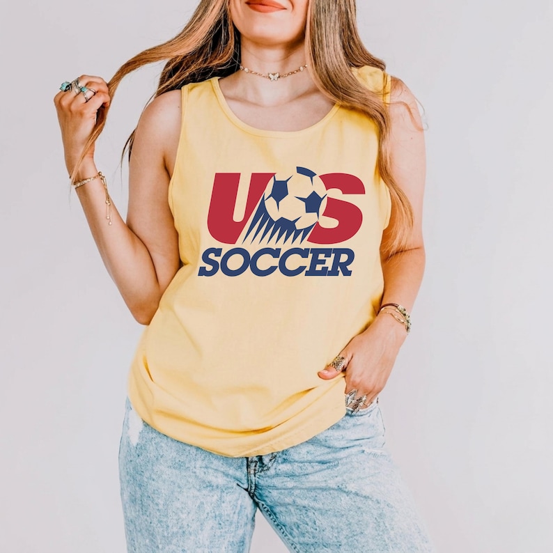 May include: A yellow sleeveless tank top featuring a graphic design with the red letters US, a white and blue soccer ball replacing the letter S, and the word SOCCER printed in blue underneath. The design is centered on the front of the shirt.