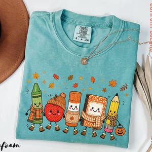 May include: A seafoam green short-sleeve t-shirt with a crew neck and a Comfort Colors label. The graphic features five anthropomorphic school supplies including a crayon, apple, glue stick, notebook, and pencil, all wearing autumn-themed accessories.