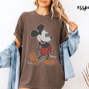 May include: Brown t-shirt featuring a classic Mickey Mouse graphic. The cartoon character is depicted in black, white, red, and brown. The word "Disney" is printed below the character. The word "esspresso" is printed in the upper right corner.