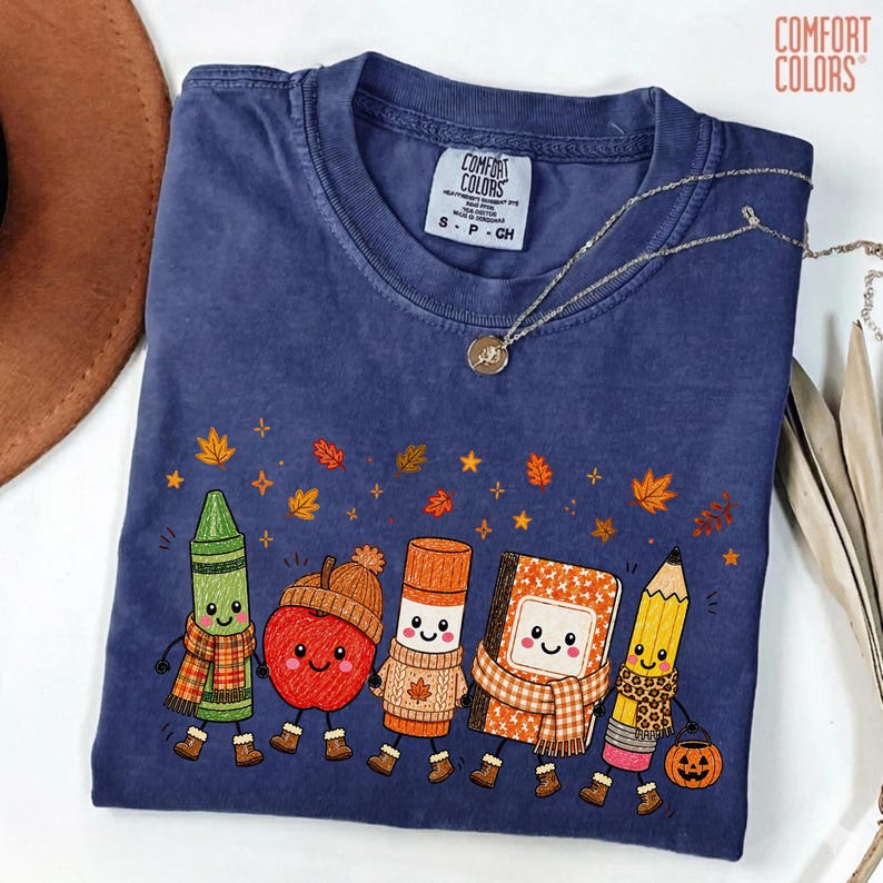 May include: A navy blue short-sleeve t-shirt featuring a graphic of a green crayon, a red apple, an orange glue stick, a notebook, and a yellow pencil, all with faces and autumn accessories. The shirt has a Comfort Colors label at the neck.