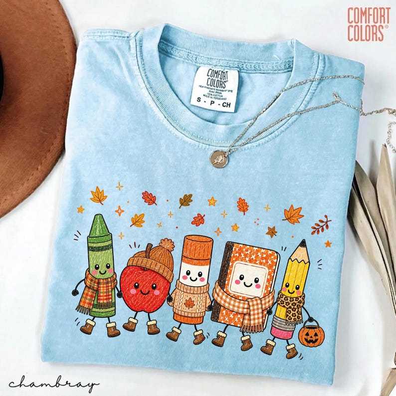May include: A light blue Comfort Colors t-shirt featuring a graphic of five anthropomorphic school supplies including a green crayon, a red apple, a glue stick, a notebook, and a yellow pencil, all wearing autumn-themed scarves and boots.