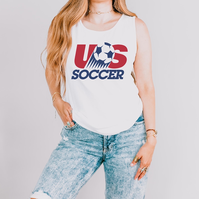 May include: A white sleeveless tank top featuring a graphic print with the red letters US, a black and white soccer ball, and the word SOCCER in dark blue text. The design is centered on the chest of the garment, which is paired with light blue acid-wash denim jeans.