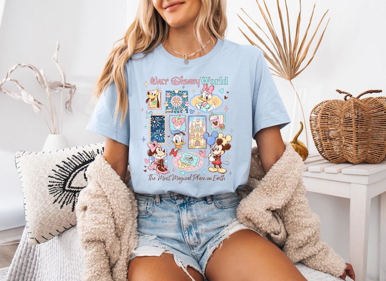 K&ouml;nnte beinhalten: Hellblaues T-Shirt mit einer farbenfrohen Grafik von Disney-Figuren und -Attraktionen. Das Design umfasst Micky Maus, Minnie Maus, Donald Duck und andere Charaktere. Der Text auf dem Shirt lautet "Walt Disney World" und "The Most Magical Place on Earth."