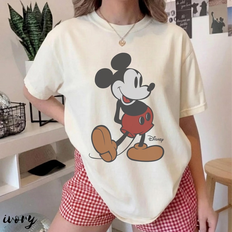 May include: Ivory-colored t-shirt featuring a large graphic of Mickey Mouse in his classic pose. Mickey is depicted with black ears, a white face, red shorts, and brown shoes. The Disney logo is printed below the character.