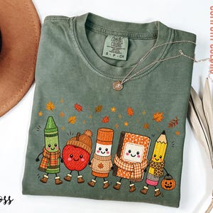 May include: A moss green short-sleeve t-shirt featuring a graphic of five anthropomorphic school supplies including a crayon, apple, glue stick, notebook, and pencil, all wearing scarves and boots, surrounded by falling autumn leaves.