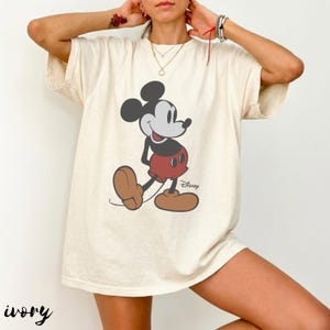 May include: Ivory-colored t-shirt featuring a classic Mickey Mouse graphic. The cartoon character is depicted in black, red, and brown, with the Disney logo visible. The word "ivory" is printed at the bottom left of the shirt.