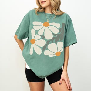 Puede incluir: Una camiseta verde de manga corta con un dise&ntilde;o floral. El gr&aacute;fico incluye grandes flores blancas con centros naranjas y tallos grises. La camiseta se usa con pantalones cortos negros.