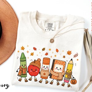 May include: An ivory t-shirt featuring a graphic of five anthropomorphic school supplies including a green crayon, an apple, a glue stick, a notebook, and a pencil, all wearing scarves and boots, surrounded by small orange and brown autumn leaves.