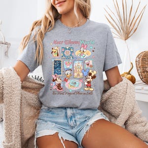 K&ouml;nnte beinhalten: Graues T-Shirt mit einer farbenfrohen Grafik von Disney-Figuren und -Attraktionen. Das Design umfasst Micky Maus, Minnie Maus, Donald Duck und andere Charaktere. Der Text auf dem Shirt lautet "Walt Disney World" und "The Most Magical Place on Earth."