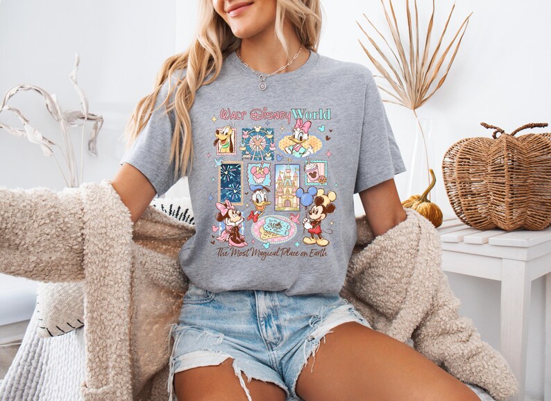 K&ouml;nnte beinhalten: Graues T-Shirt mit einer farbenfrohen Grafik von Disney-Figuren und -Attraktionen. Das Design umfasst Micky Maus, Minnie Maus, Donald Duck und andere Charaktere. Der Text auf dem Shirt lautet "Walt Disney World" und "The Most Magical Place on Earth."
