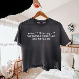 May include: A charcoal gray short-sleeve t-shirt featuring white text printed on the front that reads, your little ray of sarcastic sunshine has arrived. The garment is displayed on a wooden hanger against a bright, neutral-toned bedroom setting.