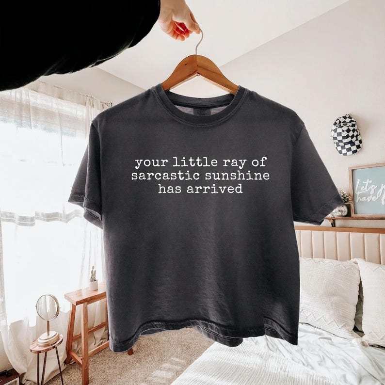 May include: A charcoal gray short-sleeve t-shirt featuring white text printed on the front that reads, your little ray of sarcastic sunshine has arrived. The garment is displayed on a wooden hanger against a bright, neutral-toned bedroom setting.