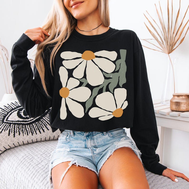 Puede incluir: Sudadera negra de manga larga con un dise&ntilde;o floral. El gr&aacute;fico incluye grandes flores blancas con centros color canela y tallos verdes. La sudadera se combina con pantalones cortos de mezclilla.