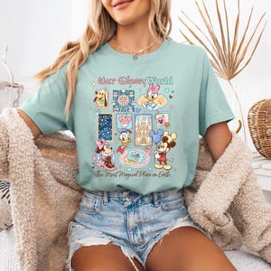 K&ouml;nnte beinhalten: Hellblaues T-Shirt mit einer farbenfrohen Grafik mit Disney-Figuren und dem Text "Our Disney World, the Most Magical Place on Earth". Das Design enth&auml;lt Bilder von Mickey Mouse, Minnie Mouse und anderen Figuren.