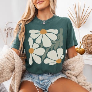 Puede incluir: Una camiseta verde azulado con un dise&ntilde;o floral de grandes margaritas blancas con centros amarillos y hojas verdes. La camiseta se combina con pantalones cortos de mezclilla y una rebeca color crema.