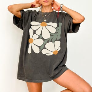 Puede incluir: Camiseta gris oscuro con un dise&ntilde;o floral grande. El gr&aacute;fico muestra tres margaritas blancas con centros naranjas y hojas verdes. La camiseta tiene un corte holgado y mangas cortas.