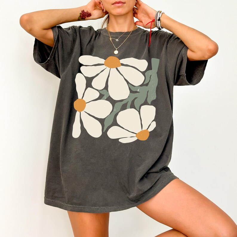 Puede incluir: Camiseta gris oscuro con un dise&ntilde;o floral grande. El gr&aacute;fico muestra tres margaritas blancas con centros naranjas y hojas verdes. La camiseta tiene un corte holgado y mangas cortas.