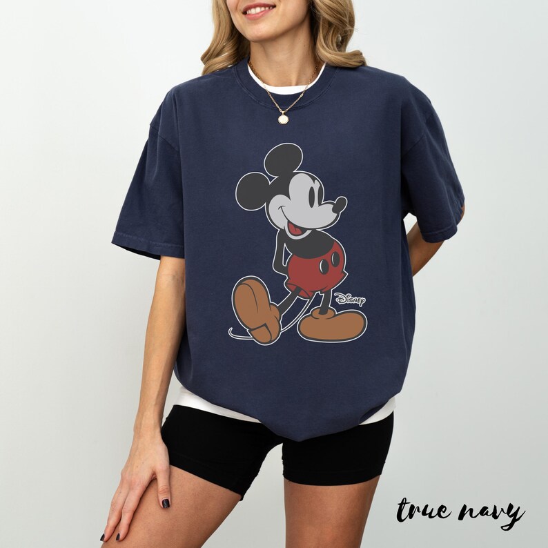 May include: Navy blue t-shirt featuring a classic Mickey Mouse graphic. The cartoon character is depicted in his iconic pose, with red shorts, brown shoes, and a smiling expression. The word "Disney" is printed below the character. The text "true navy" is at the bottom.