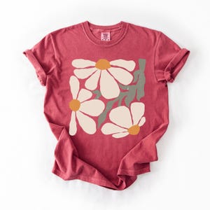 Puede incluir: Una camiseta roja con un dise&ntilde;o floral. El gr&aacute;fico incluye grandes flores blanquecinas con centros naranjas y tallos verdes. La camiseta tiene cuello redondo y mangas enrolladas, y una peque&ntilde;a etiqueta es visible cerca del cuello.