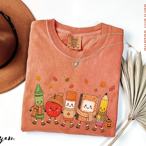 May include: A terracotta-colored crewneck t-shirt featuring a graphic of five anthropomorphic school supplies including a crayon, apple, glue stick, notebook, and pencil, all wearing autumn-themed accessories and surrounded by falling leaves.