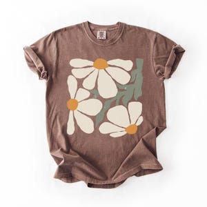 Puede incluir: Una camiseta marr&oacute;n con un dise&ntilde;o floral. El gr&aacute;fico incluye tres grandes margaritas blancas con centros naranjas y hojas verdes. La camiseta tiene cuello redondo y mangas cortas.