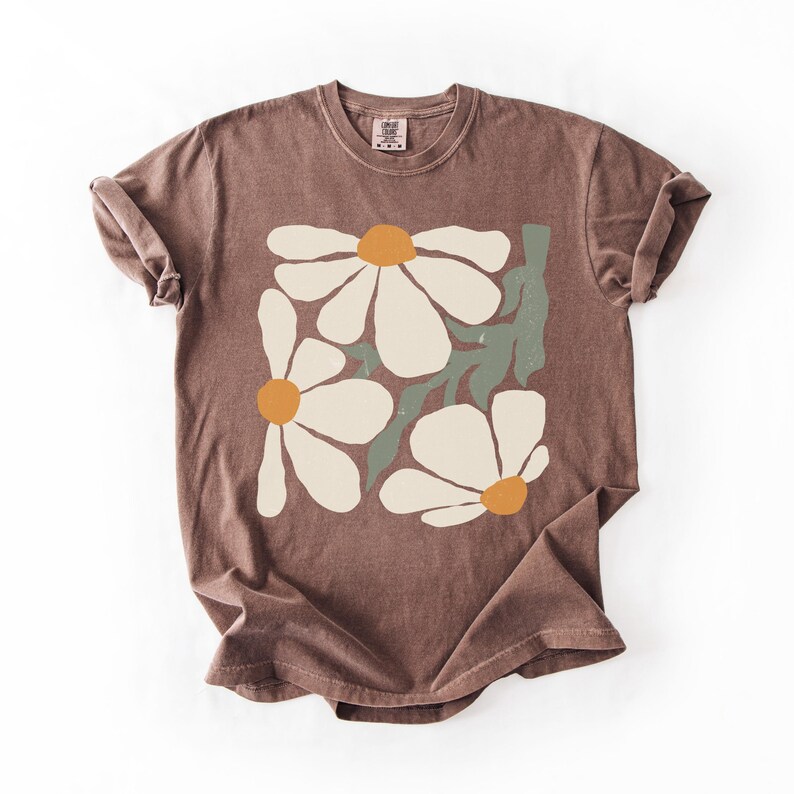 Puede incluir: Una camiseta marr&oacute;n con un dise&ntilde;o floral. El gr&aacute;fico incluye tres grandes margaritas blancas con centros naranjas y hojas verdes. La camiseta tiene cuello redondo y mangas cortas.