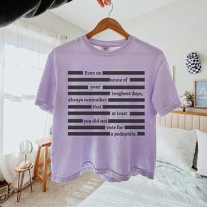 Puede incluir: Una camiseta de color lila claro con el texto "Even on some of your toughest days, always remember that at least you did not vote for a pedophile." La camiseta est&aacute; colgada en una percha de madera.