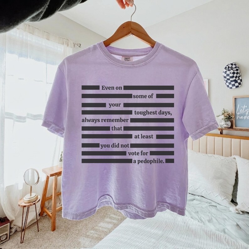 Puede incluir: Una camiseta de color lila claro con el texto "Even on some of your toughest days, always remember that at least you did not vote for a pedophile." La camiseta est&aacute; colgada en una percha de madera.